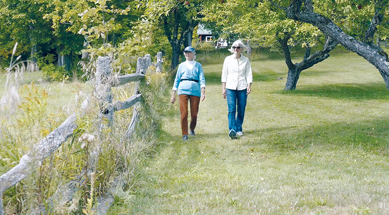 Ruth Vaill and her friend Cathy Johnston take a walk in a grassy field with trees.
